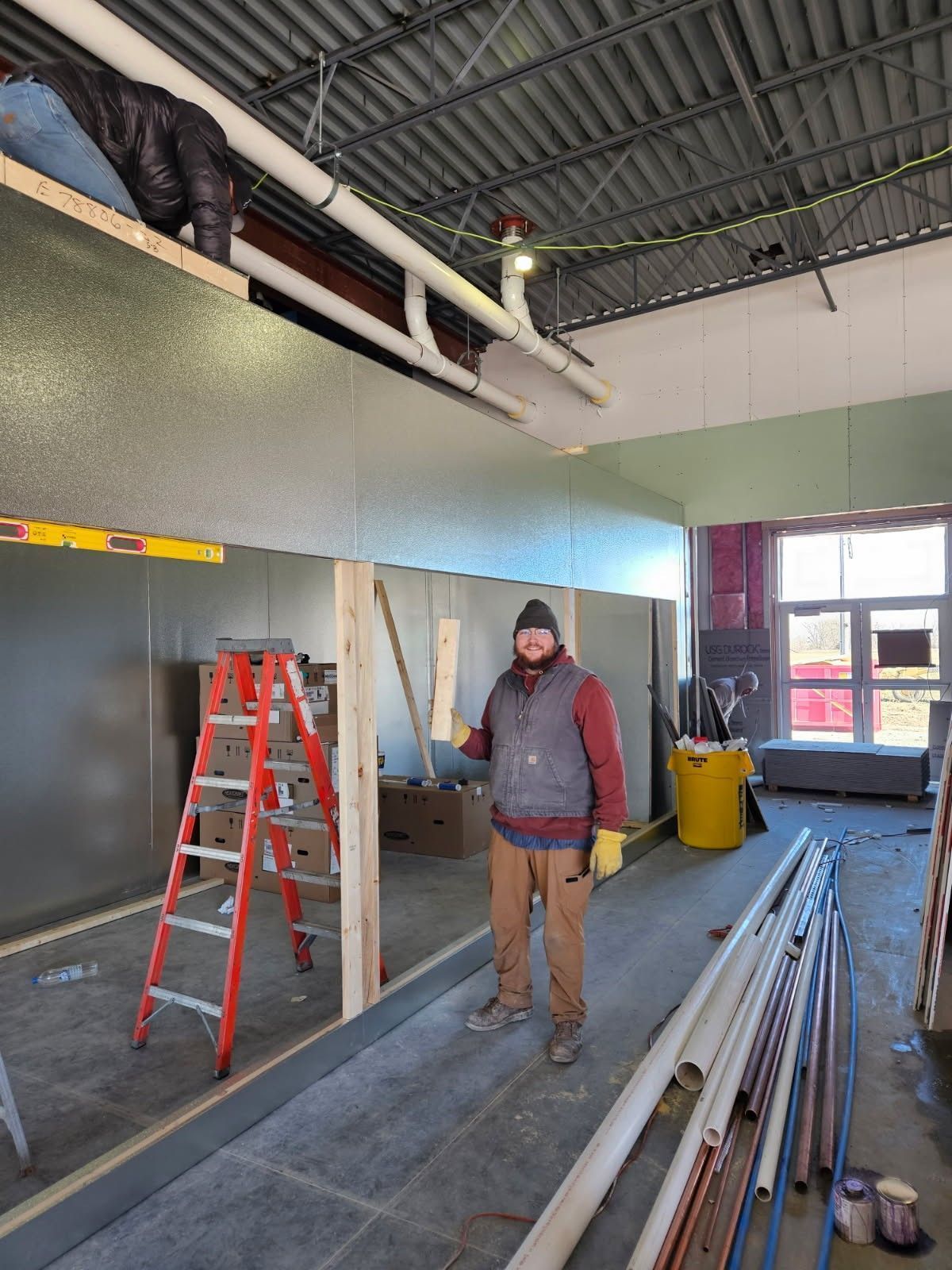 Person standing in an unfinished room with a red ladder, stacked pipes, and exposed ceiling beams.