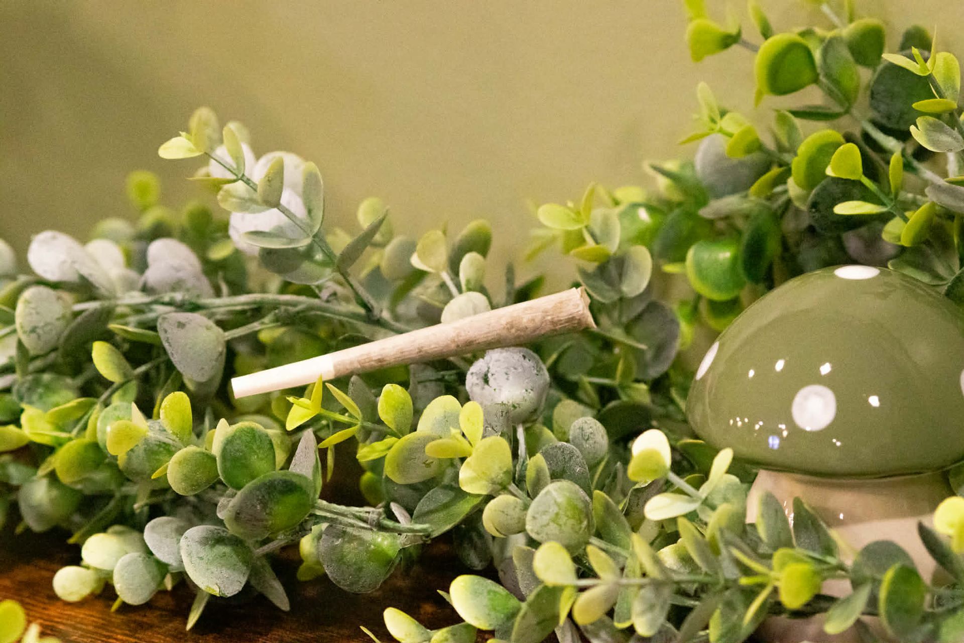 A rolled cigarette rests on a green leafy garland next to a small, sage-green ceramic mushroom figurine.