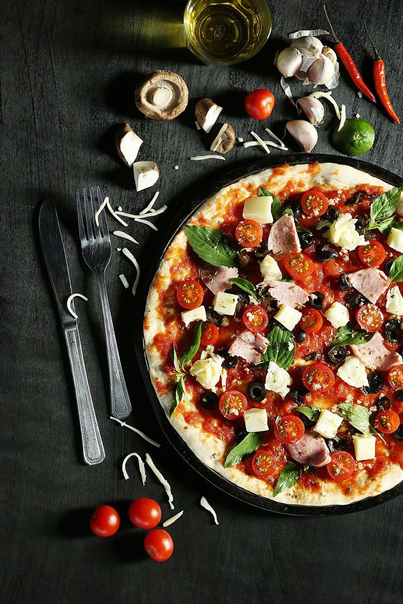 A pizza with tomatoes , olives , ham and cheese on a black plate on a table.