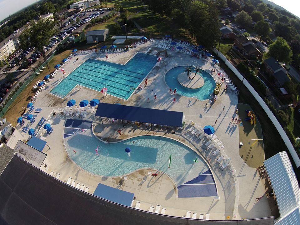 Aerial view of a public swimming pool complex with multiple pools, a lazy river, and shaded areas.