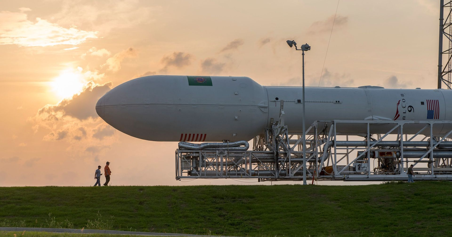 Rocket on launch pad at sunset with two people standing nearby, grassy foreground and orange sky
