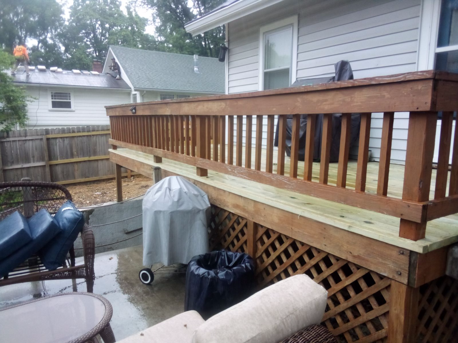 Newly built wooden deck with light colored deck and darker brown stained railing around the deck and lattice skirting beside a house, with covered grill and yard items below.