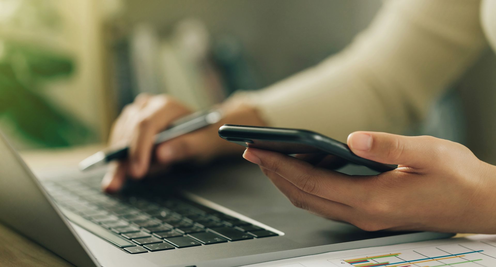 Person using a laptop and smartphone simultaneously, holding a pen.