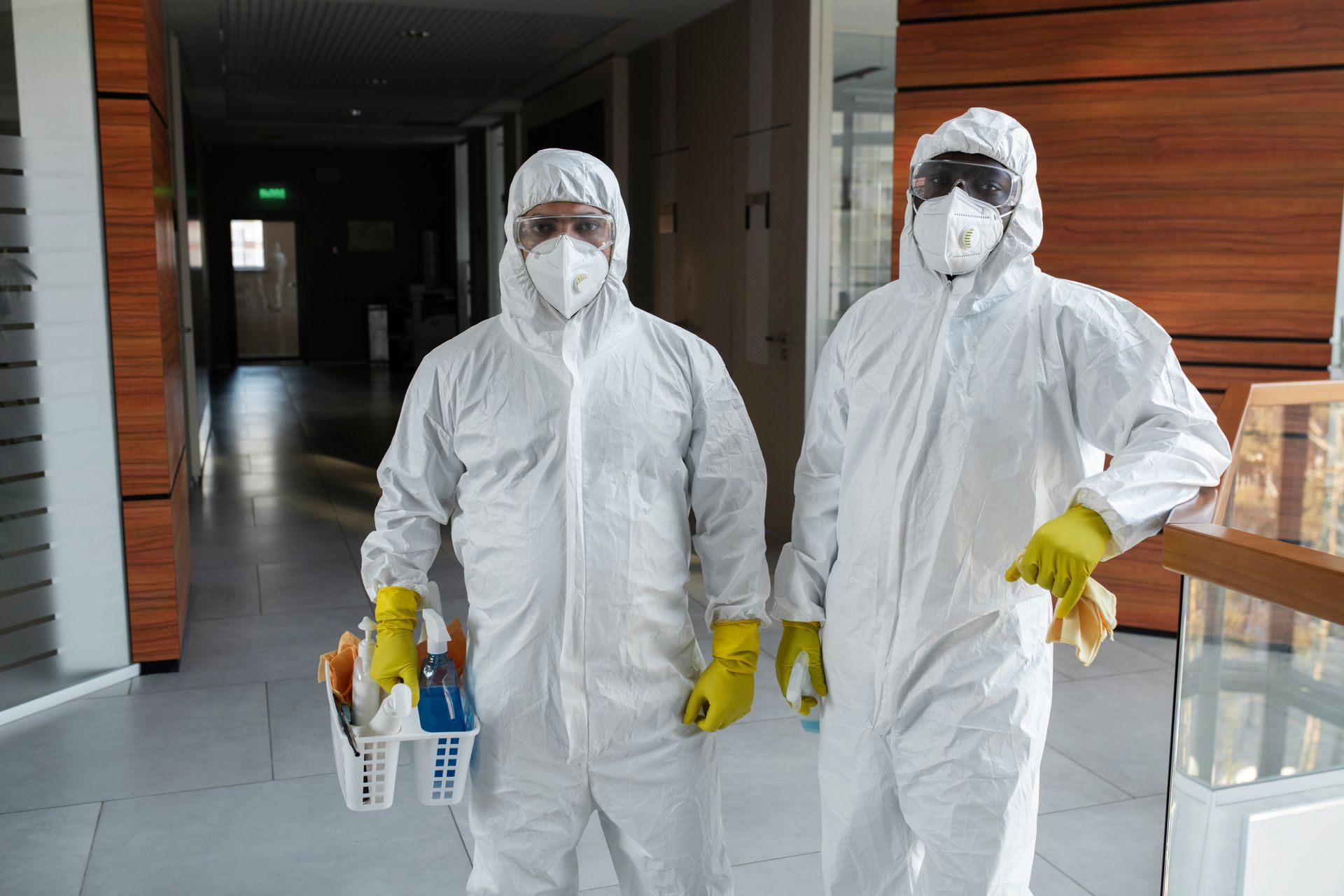 Two people in white hazmat suits, masks, and yellow gloves standing in a building hallway, one holding cleaning supplies.