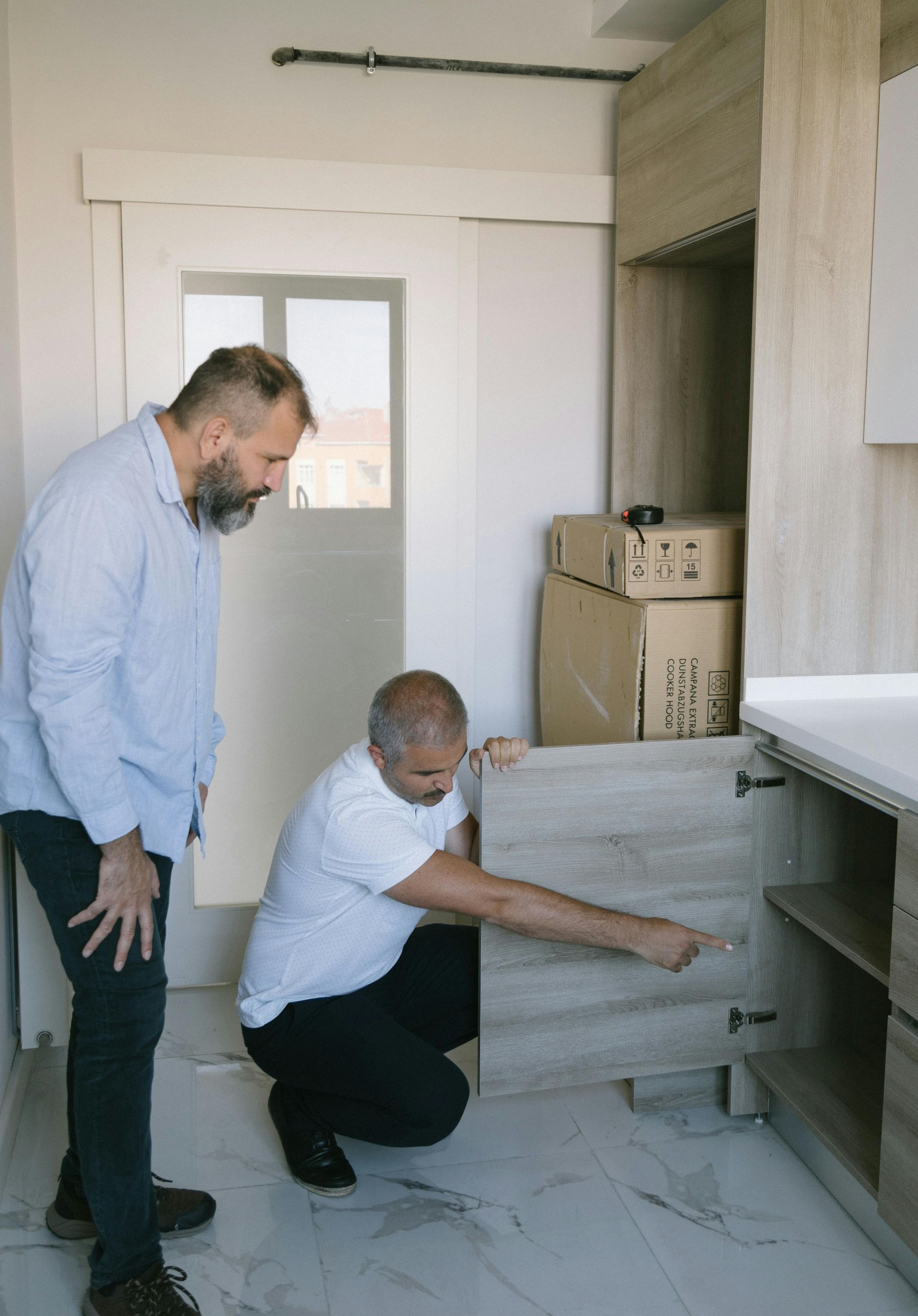 Two men examine kitchen cabinet; one points inside. Boxes in background. Light wood cabinets, tile floor.