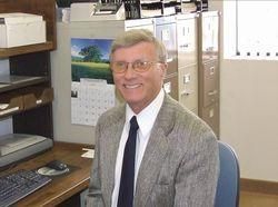 Man in a suit smiles at a desk with a computer, filing cabinets in the background.