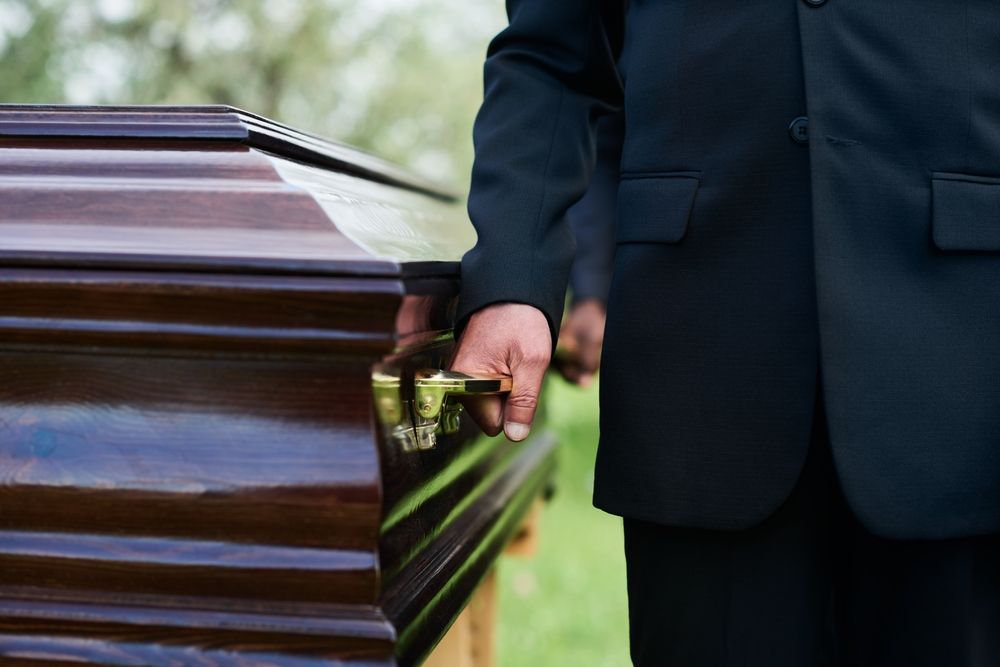 Person in black suit holding a brown coffin handle outdoors on a green lawn.