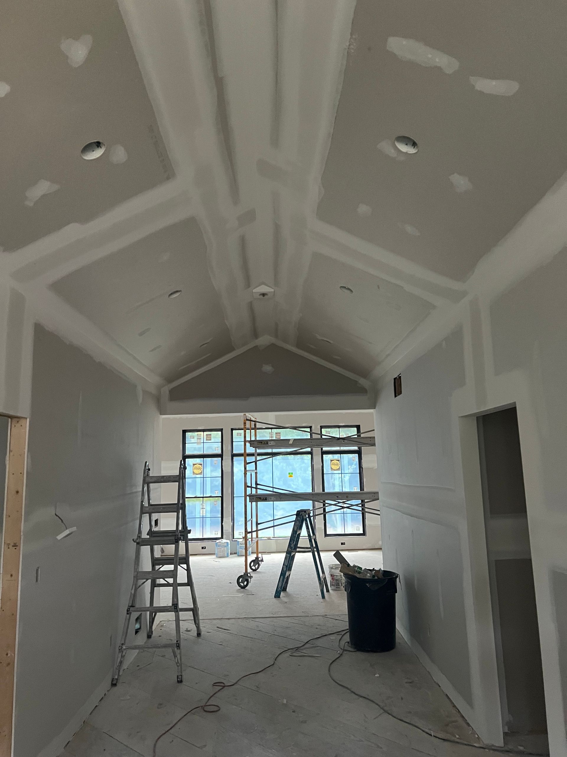 Interior of a room under construction with drywall, ladders, tools, and a window at the far end