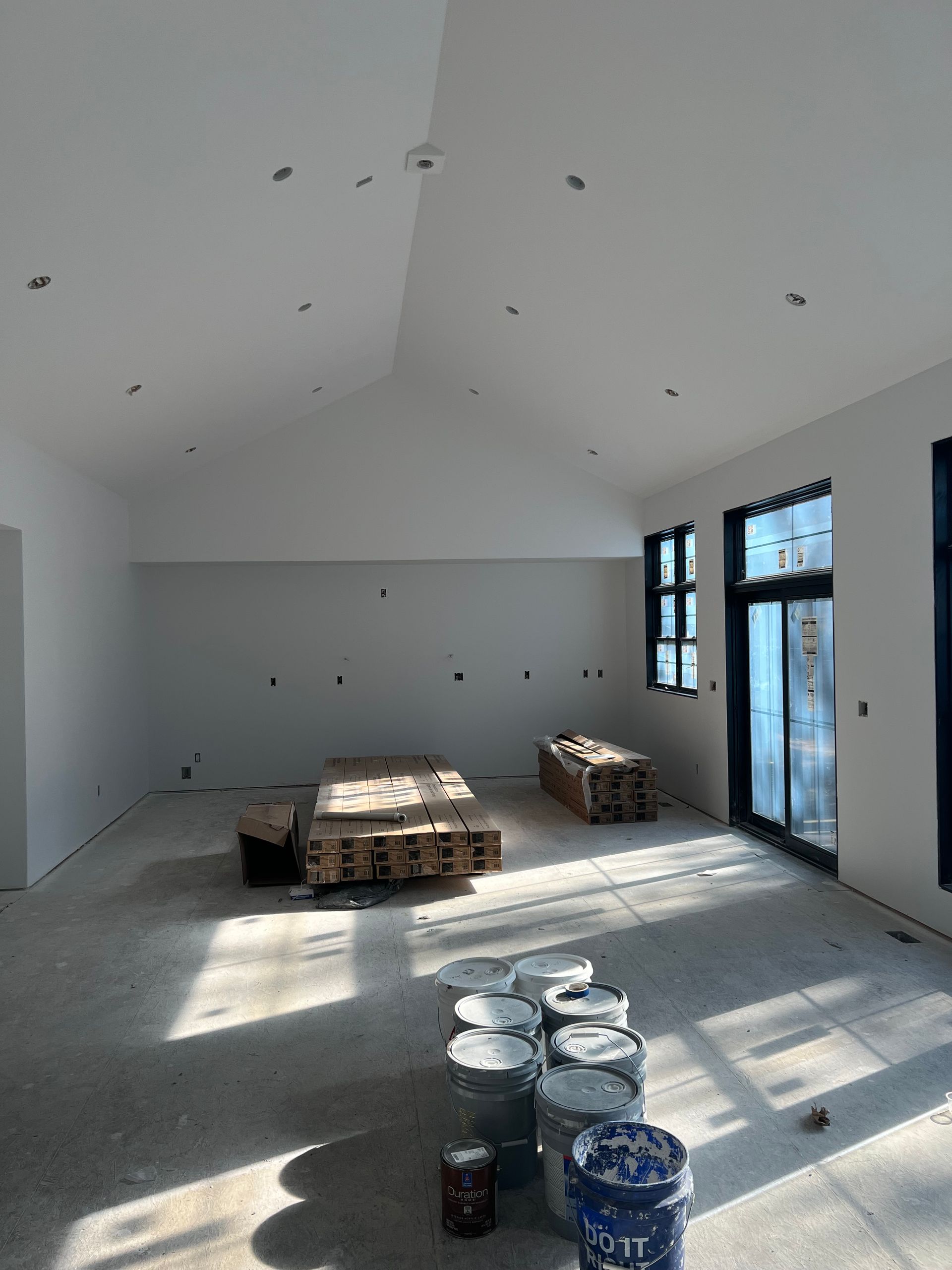 Empty room under construction with white walls, pallets, paint cans, and sunlight through windows and a glass door