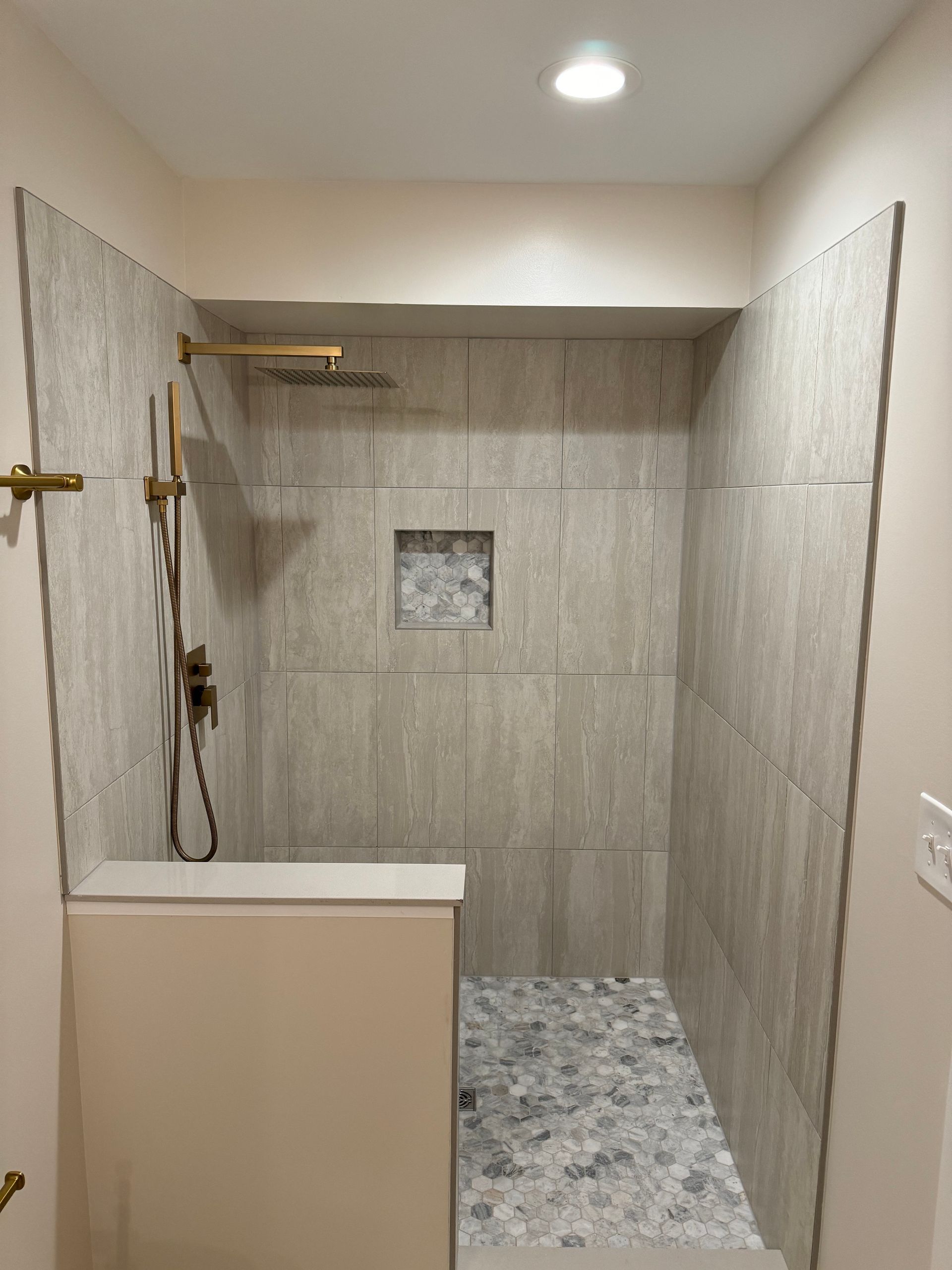 Empty tiled shower stall with a bench, brass fixtures, and a recessed niche under a ceiling light