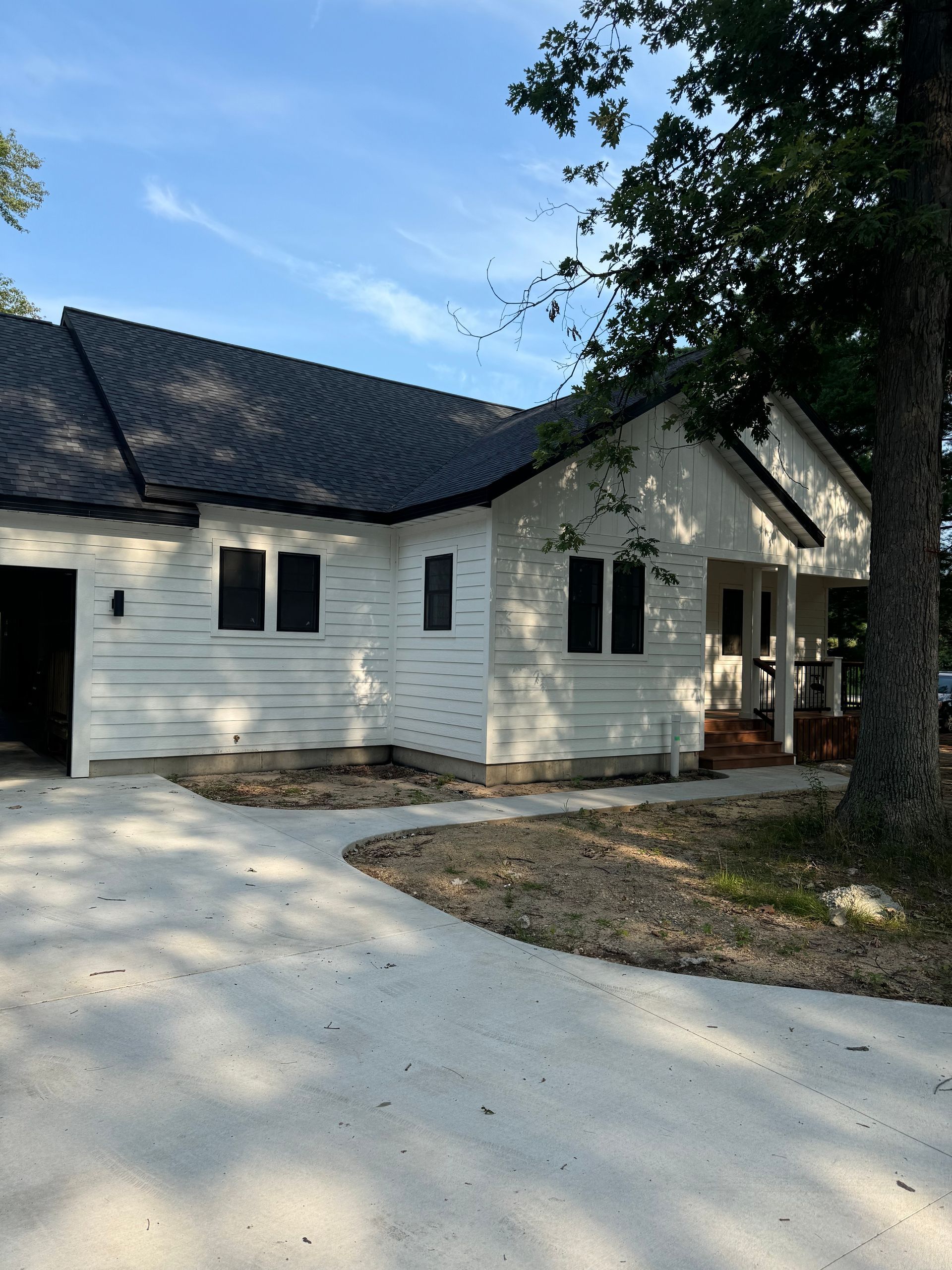 White house with dark roof, front porch, and large tree beside a curved driveway under a blue sky.