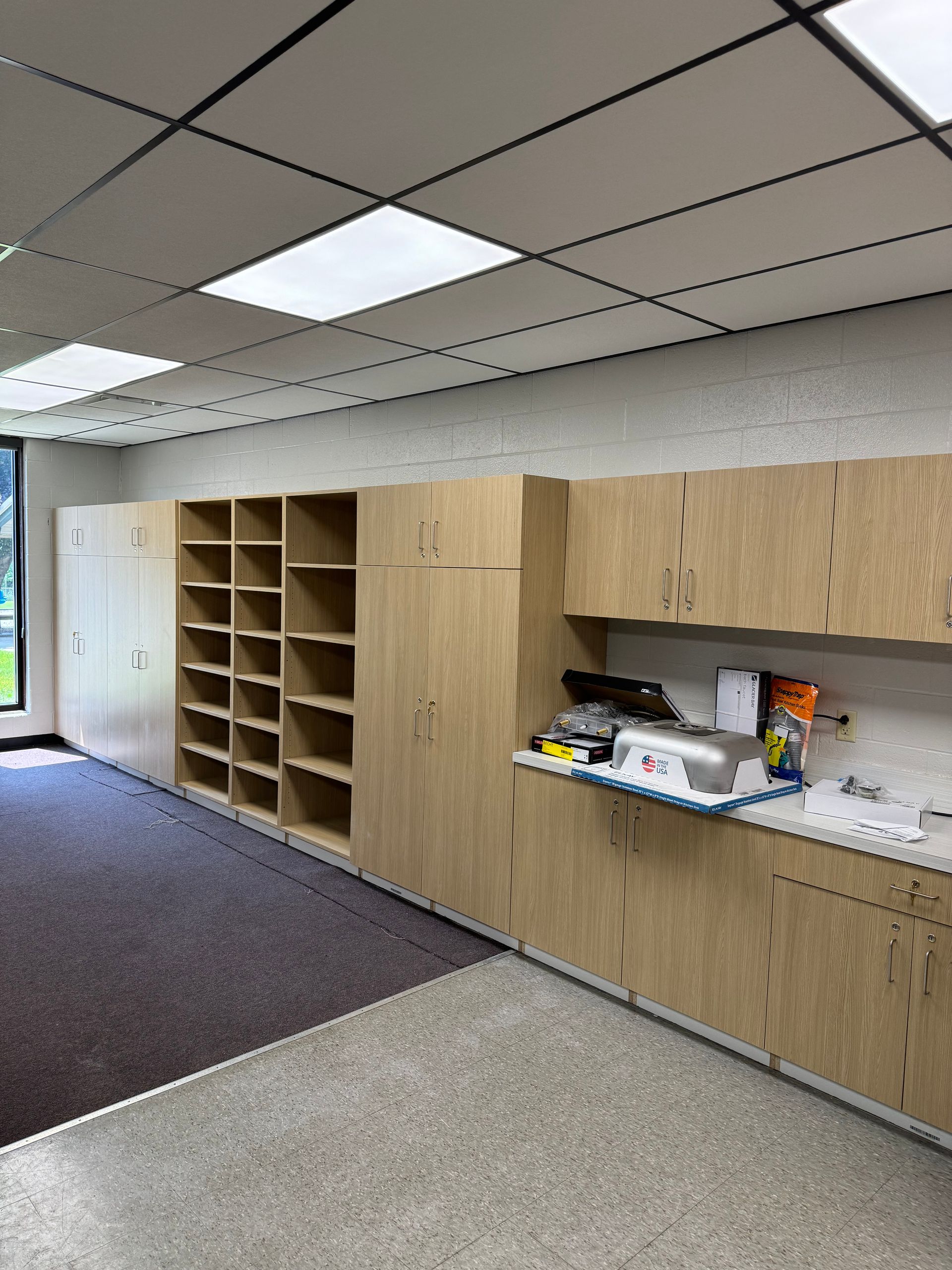 Office break room with wood cabinets, open mail slots, and countertop supplies under fluorescent ceiling lights