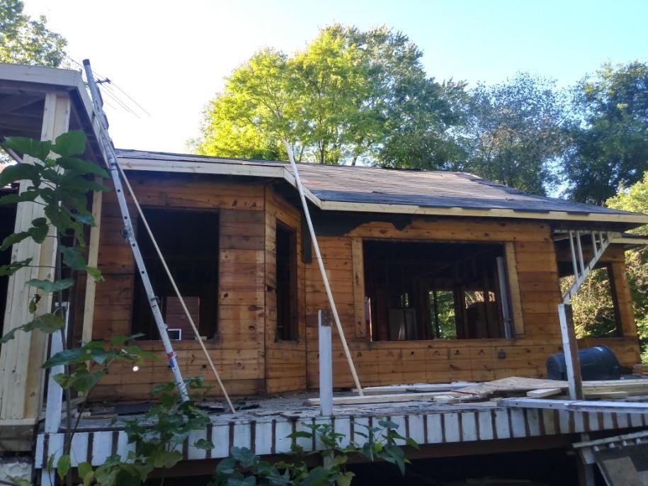 Wooden cabin under construction with a ladder, porch, and trees in the background