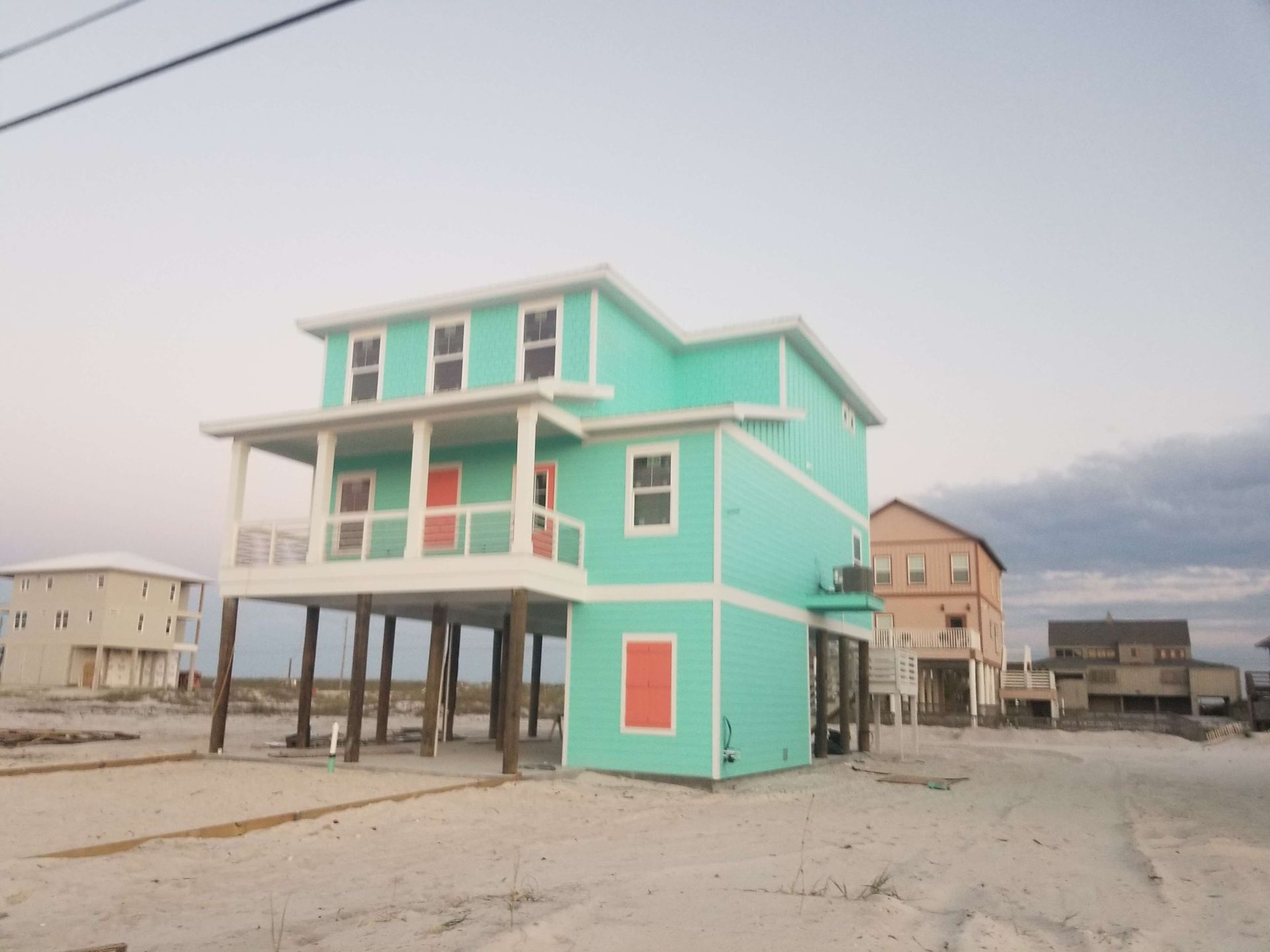 A two-story turquoise beach house on stilts, with orange doors, on a sandy lot.