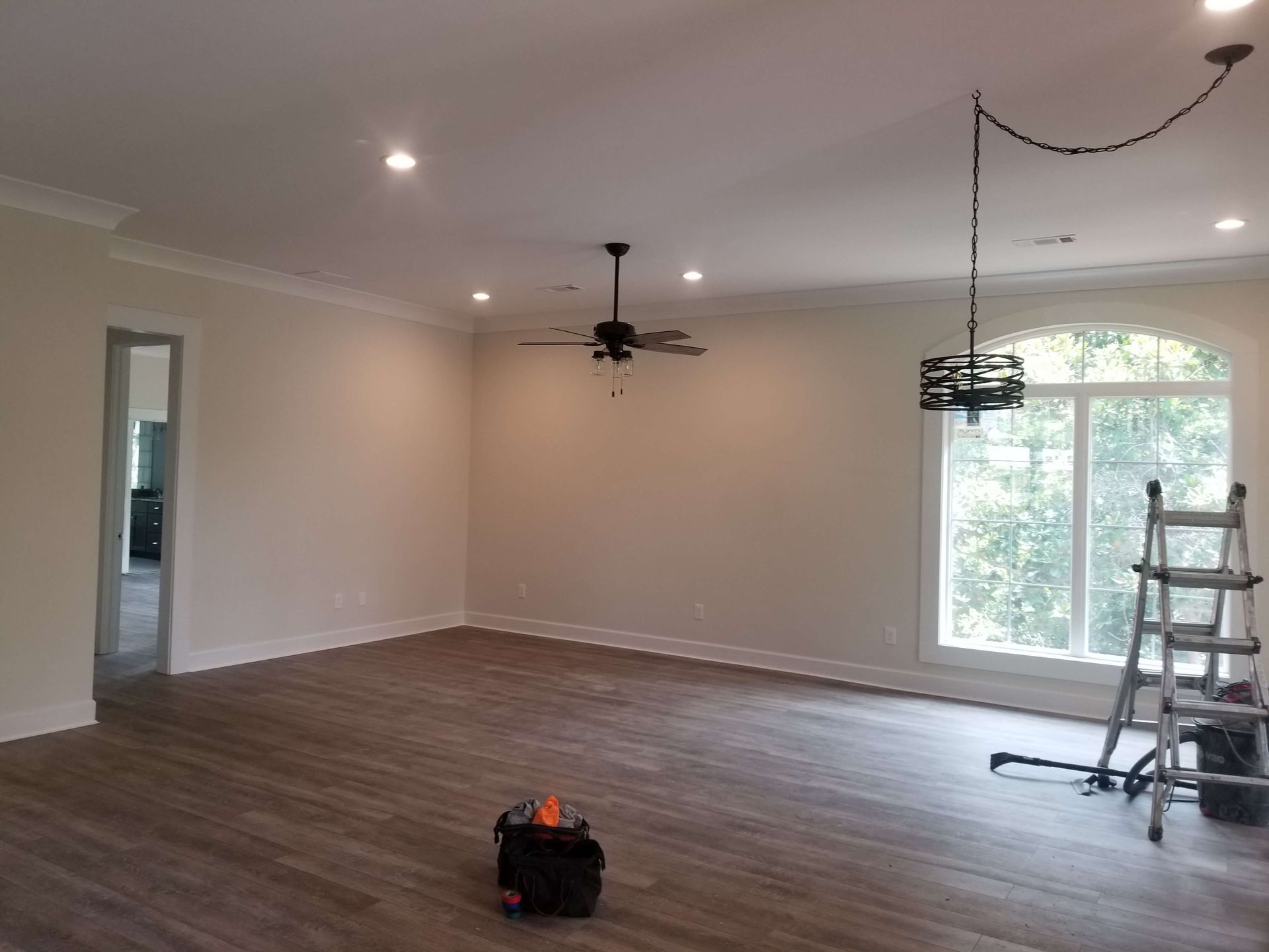 Spacious, freshly painted room with hardwood floors, a ceiling fan, and an arched window. A ladder sits near the window.