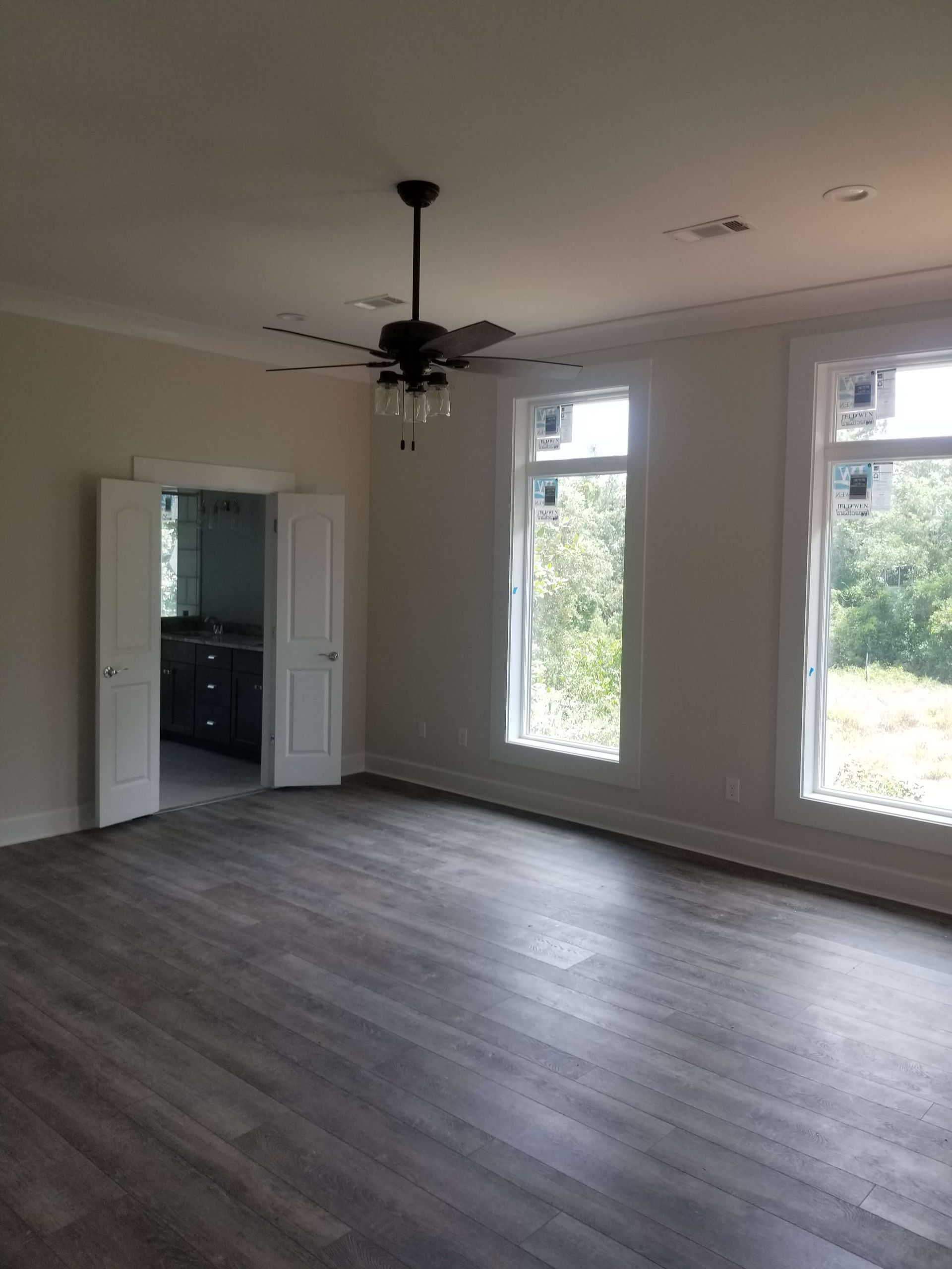 Empty room with gray floors, white walls, three windows, a ceiling fan, and a doorway to a bathroom.