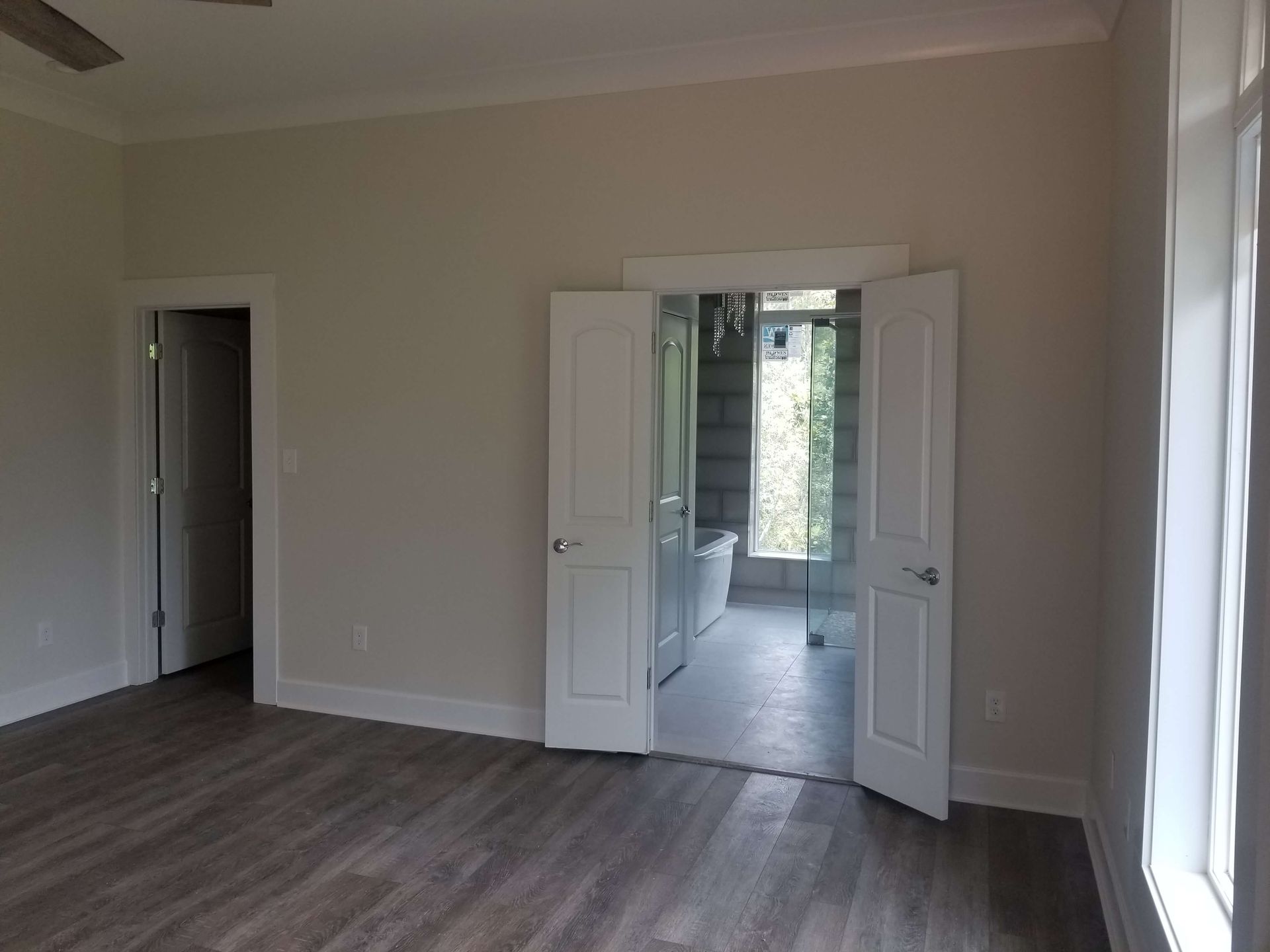 Bedroom with open doors to a bathroom. Gray floors, white doors/trim, neutral walls, natural light from a window.