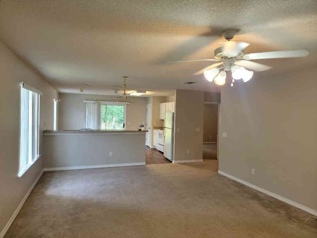 Open-concept living space with tan walls, carpet, and a ceiling fan; partially visible kitchen.