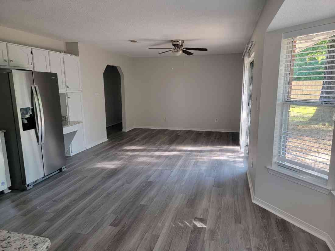 Empty living room with light gray floors and walls, white cabinets, stainless steel refrigerator, and a window.