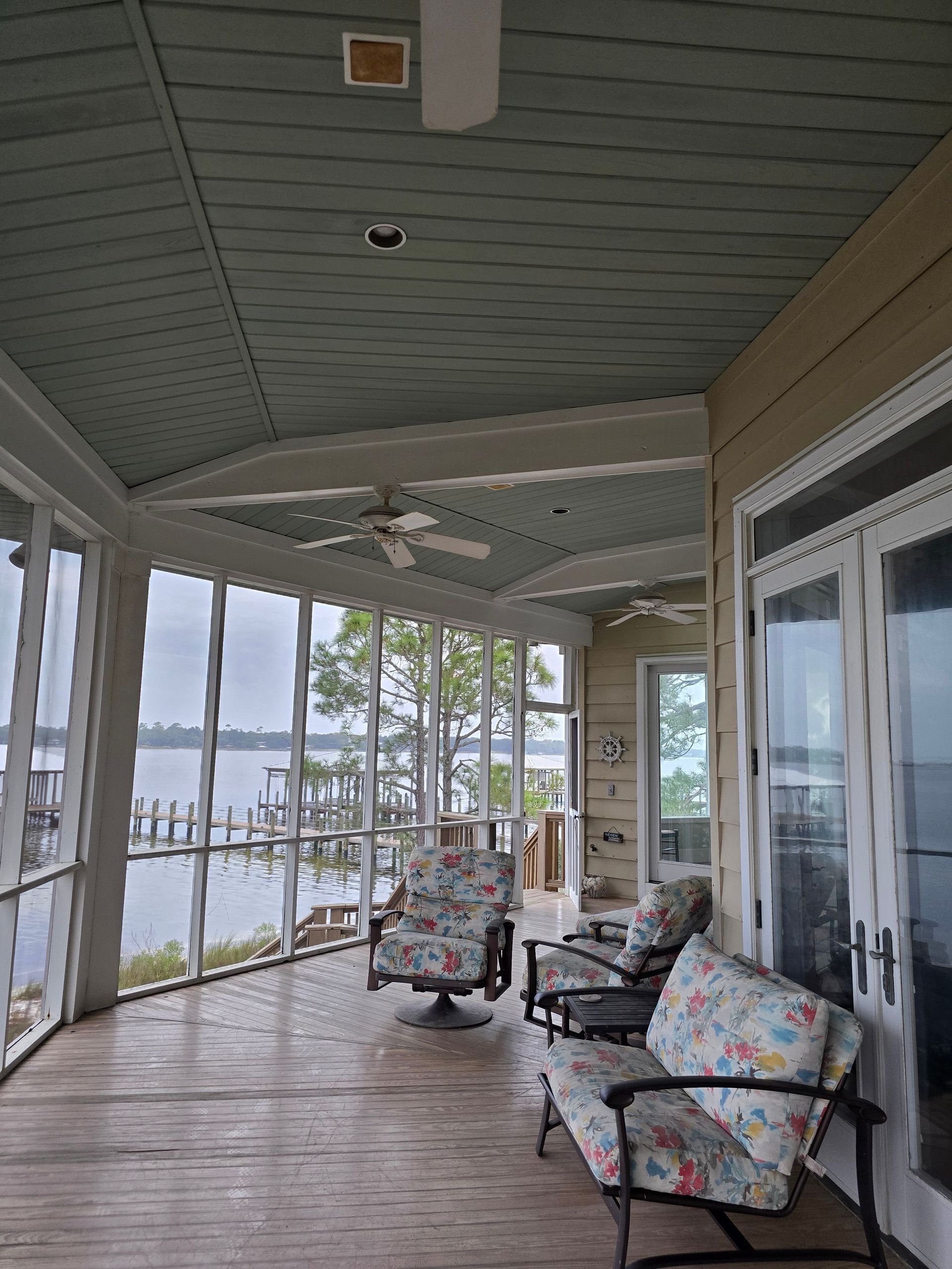 Screened porch overlooking water, furnished with chairs. Blue ceiling, wooden floor.