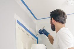 Man painting trim blue with a brush, wearing gloves and a white shirt. Blue tape on the wall.