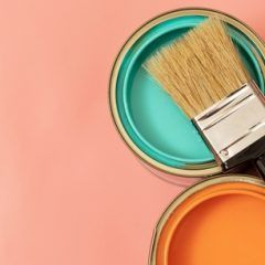 Two open paint cans, one teal and one orange, with a paintbrush resting in the teal paint; on a pink surface.