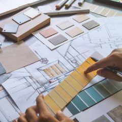 Hands pointing to a yellow paint swatch on an architectural blueprint, surrounded by design samples.