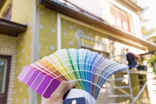 Person holding a colorful paint swatch fan in front of a house being painted.