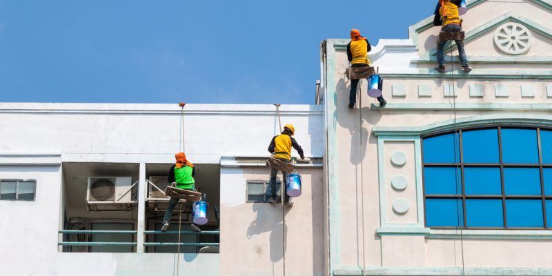 Three painters on ropes painting a building exterior, blue sky background.