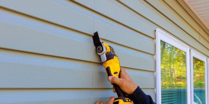A person using a yellow power tool to cut vinyl siding on a blue house.