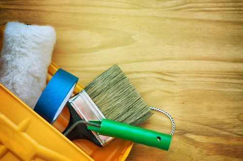 Paint supplies in a yellow tray on a wood surface, including a roller, brush, and tape.