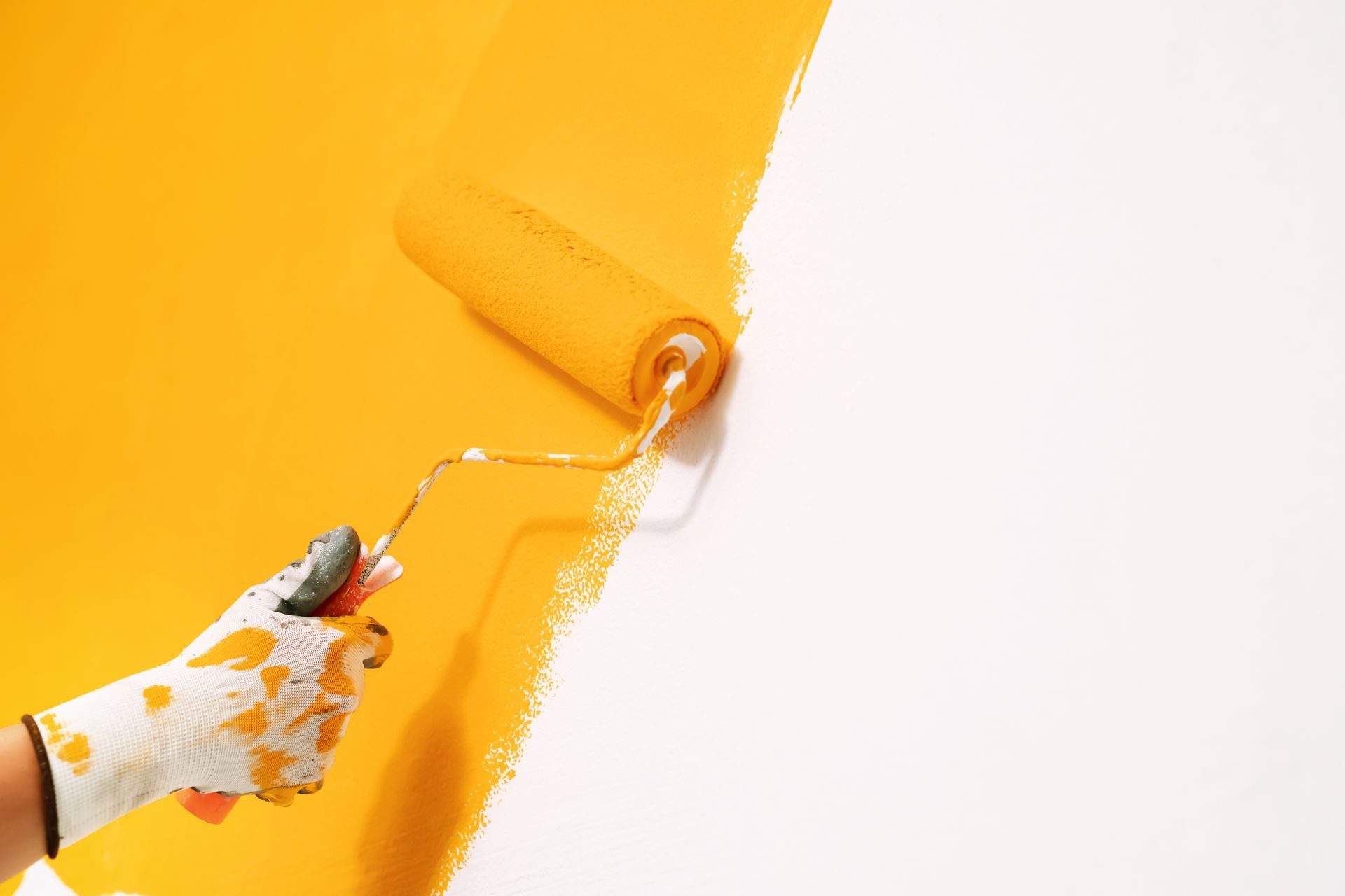 Hand in paint-splattered glove paints a white wall yellow with a roller.