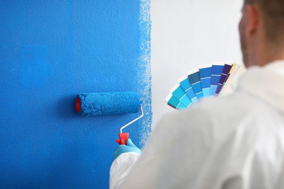 Person painting a blue wall with a roller, holding a color palette.
