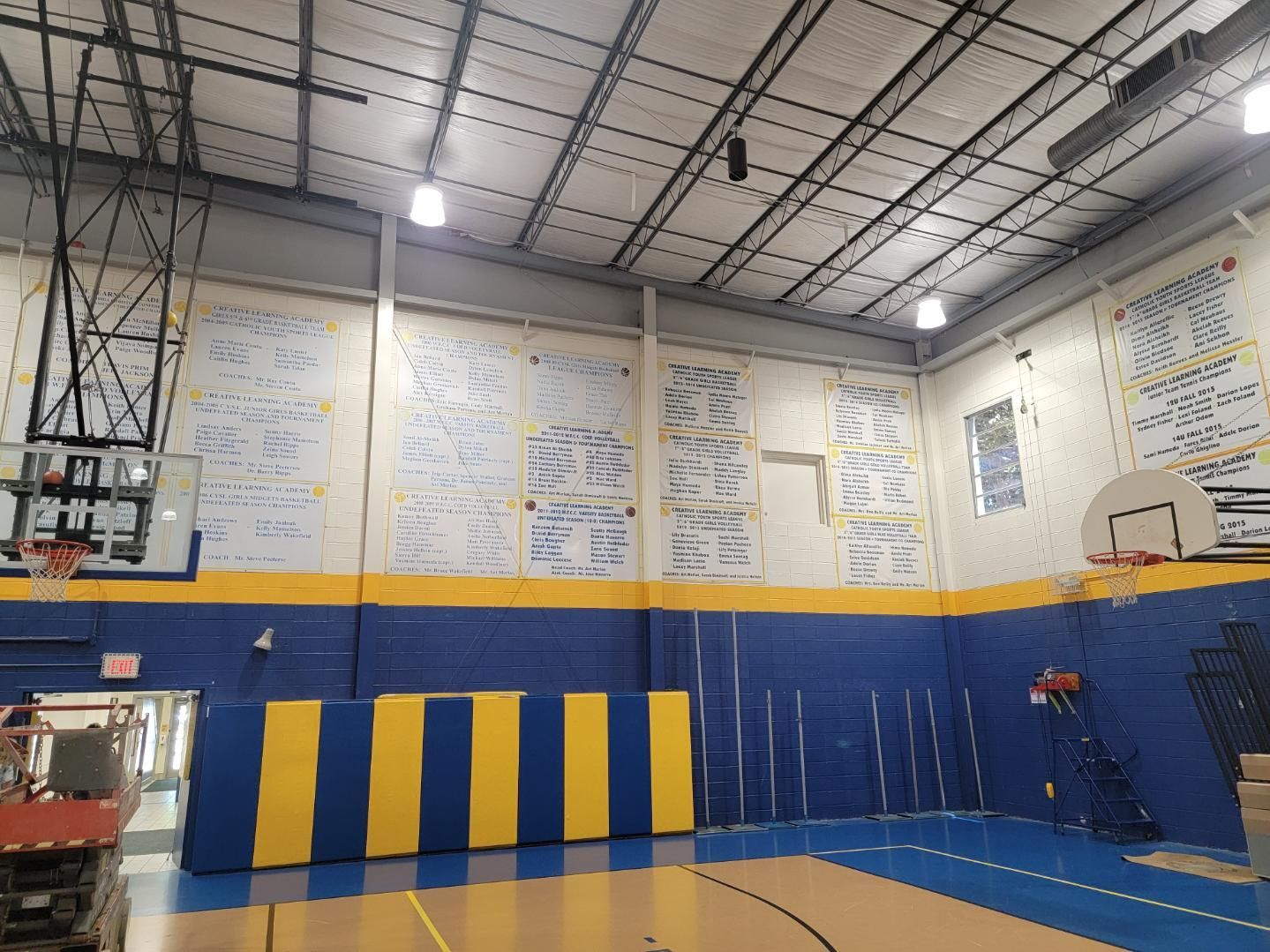 Gymnasium interior with blue and yellow padded walls, basketball hoops, and writing on the upper walls.