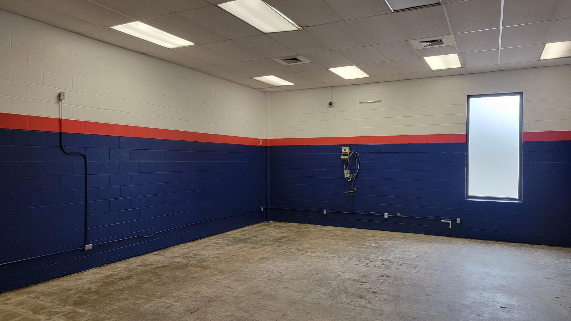 Empty room with blue and red painted walls, concrete floor, fluorescent lights, and a window.