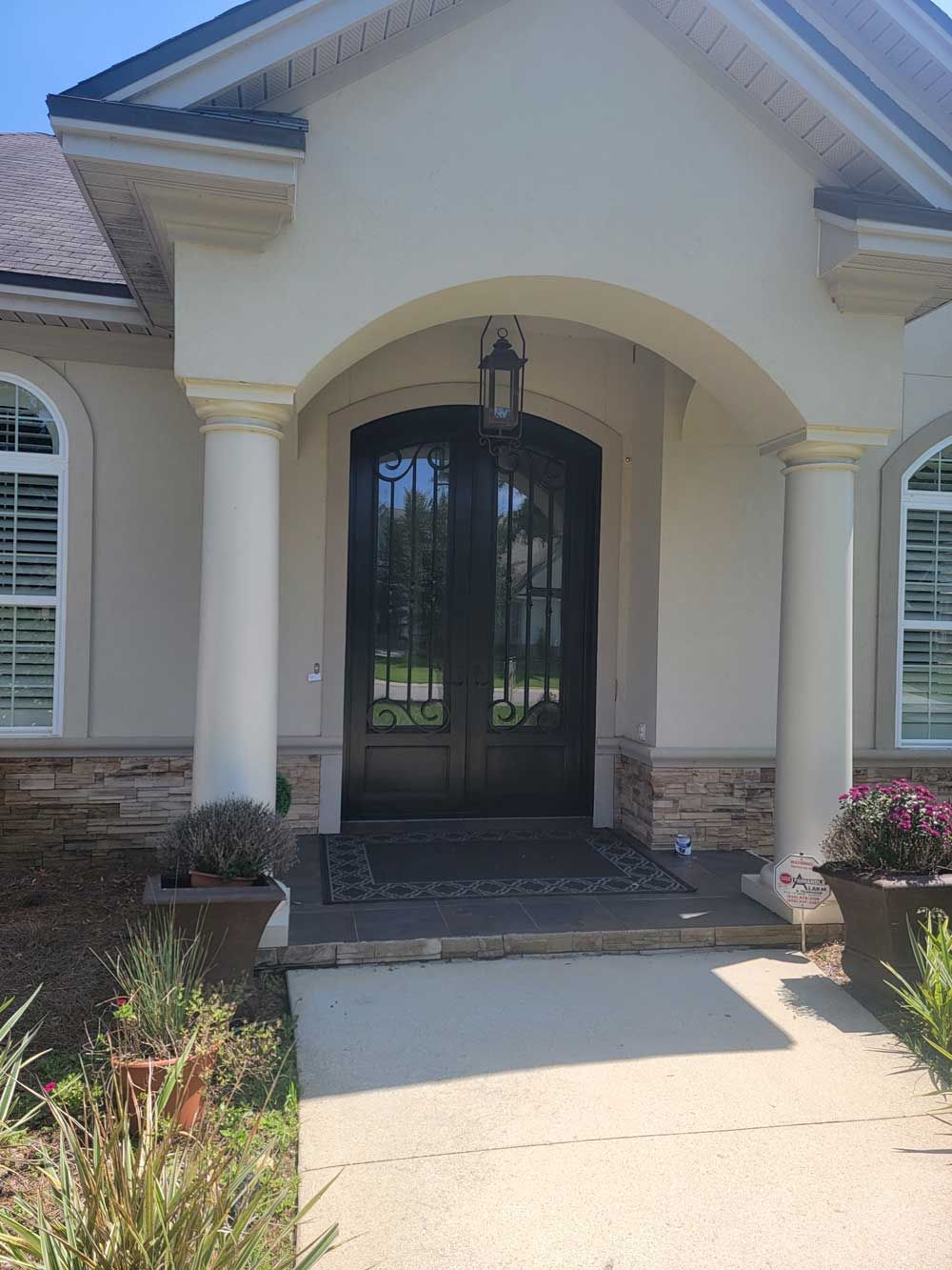 Front entrance of a house with a dark iron double door. Stone and stucco exterior, columns, and potted plants.
