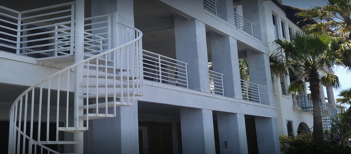A white spiral staircase on a multi-story building with balconies and palm trees.
