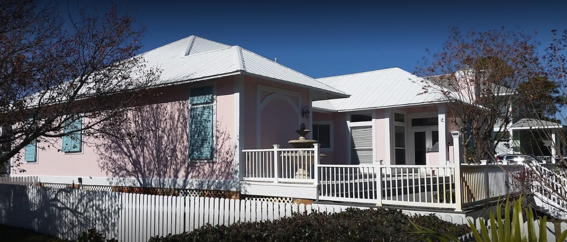 Pink house with white railing and roof, surrounded by greenery and a blue sky.