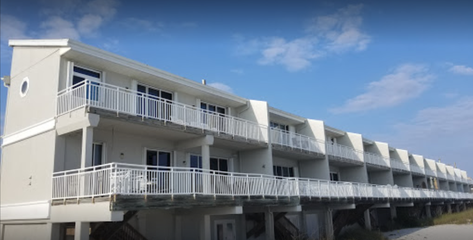 Two-story light grey beachside apartment building with white balconies under a blue sky.