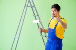 Painter in blue overalls, holding a paint roller, points at the viewer, standing by a ladder and green wall.