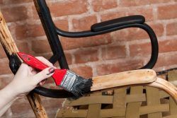 Person paints a wooden chair with a red-handled brush, against a brick wall background.
