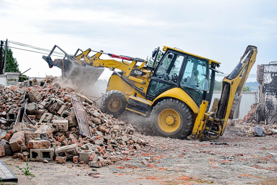 Yellow backhoe loader clearing rubble at a demolition site, with dust and broken bricks everywhere