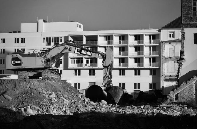 Black-and-white scene of excavators demolishing a building amid rubble in front of an apartment block