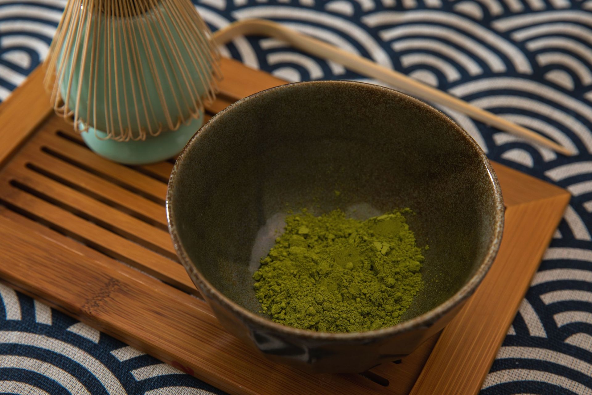 A ceramic bowl containing green matcha powder resting on a wooden tray alongside a bamboo whisk.