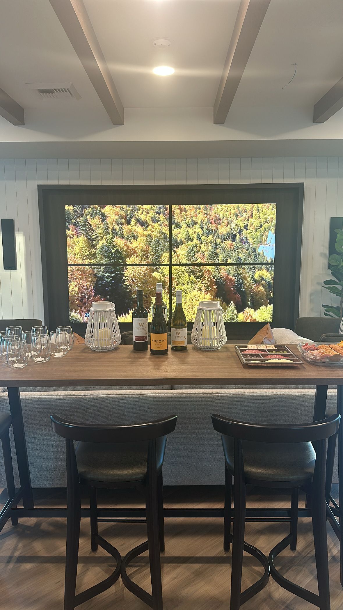 A bar area with two chairs, bottles of wine, and glasses set before a large window displaying a vibrant, autumnal scene.