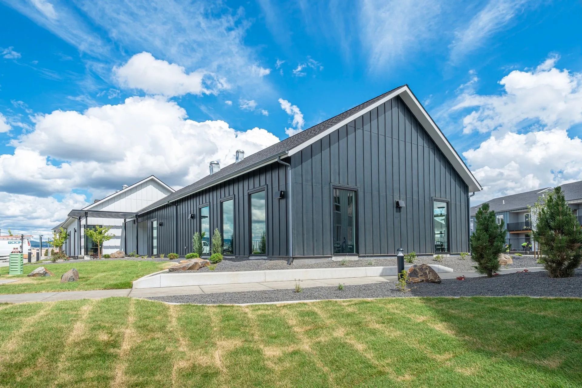 A dark grey building featuring a pitched roof and large windows standing near a concrete path and gr
