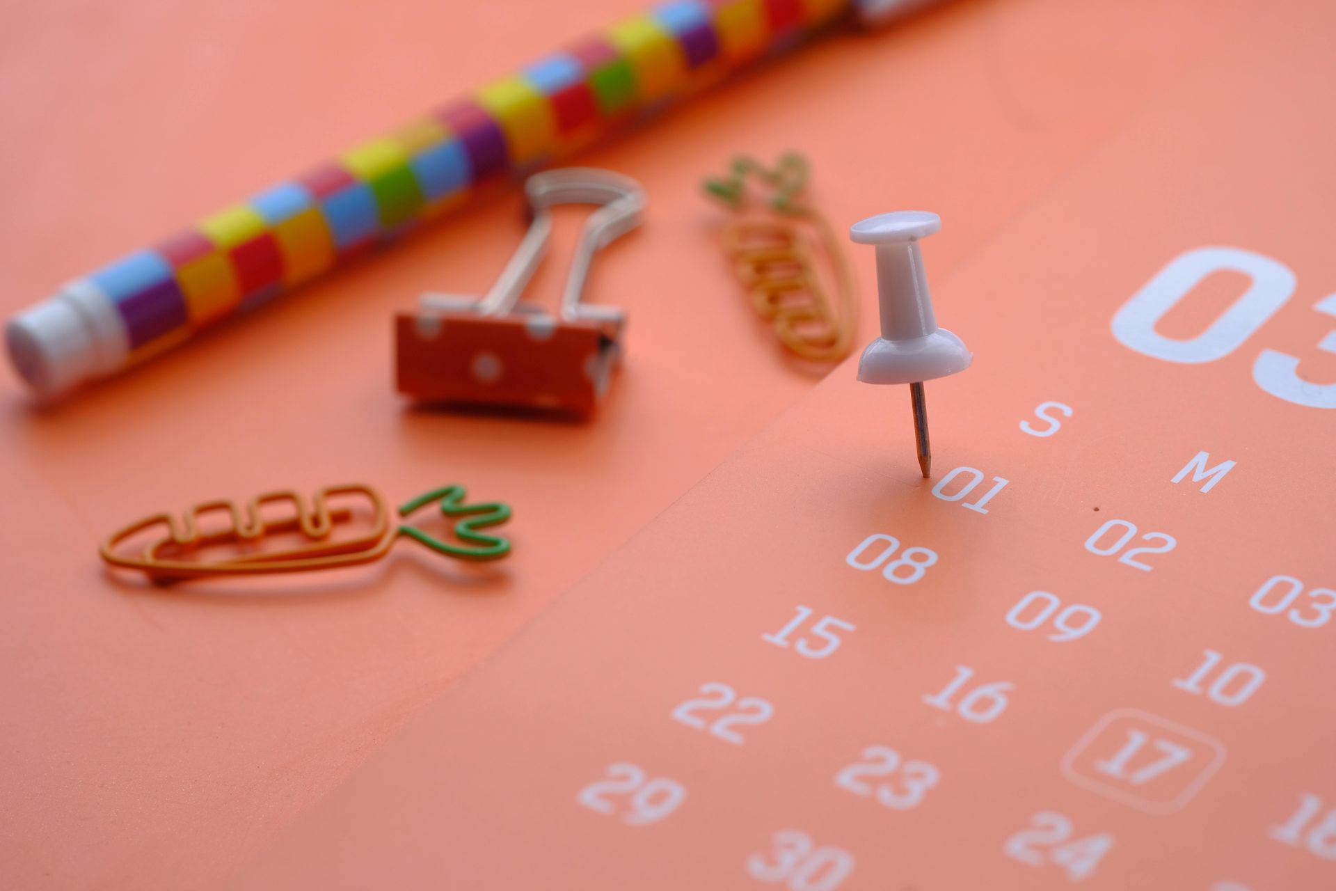 A white pushpin sticking into the first day of an orange calendar resting near colorful clips and a striped pen.