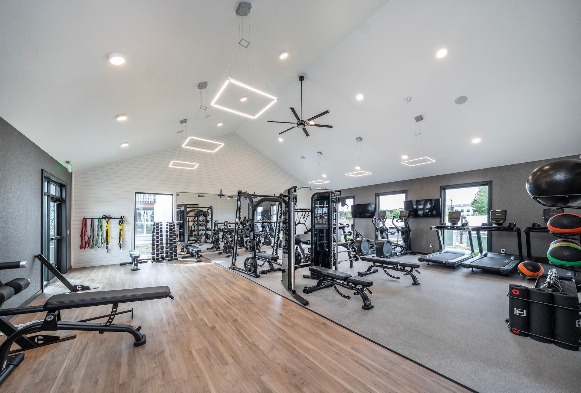 Modern gym with weight machines, benches, and treadmills; light wood floor, grey walls, and white ceiling.