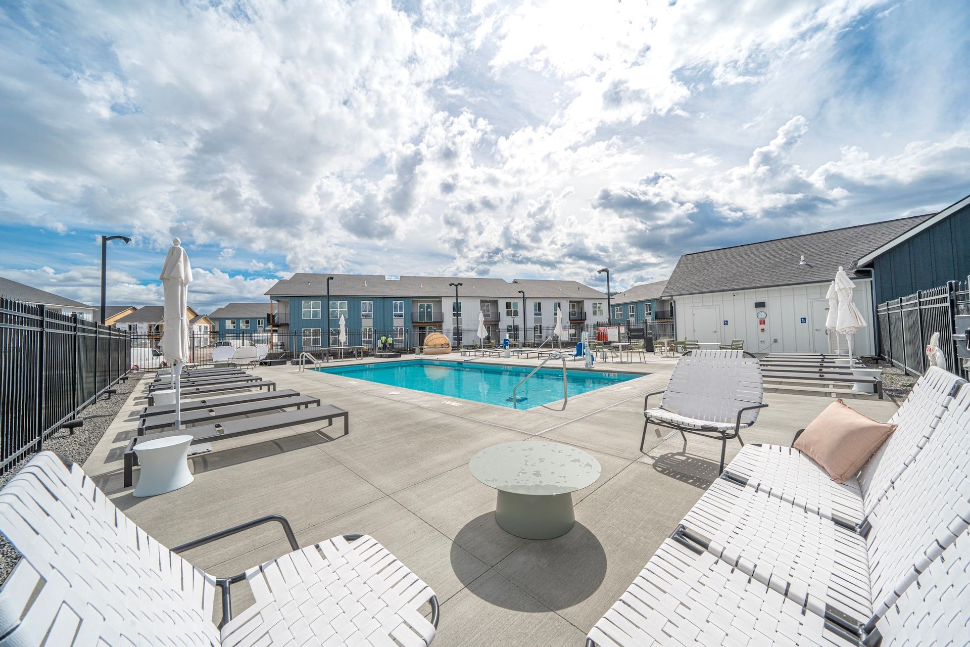 Pool area with lounge chairs, a round table, and apartment buildings on a cloudy day.