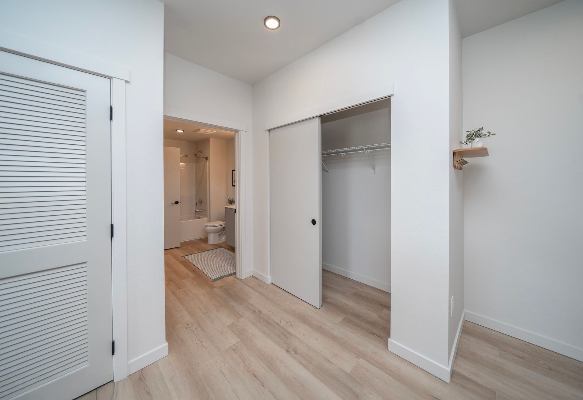 Hallway with white walls, wood-look flooring, open closet, bathroom doorway, and patterned door.